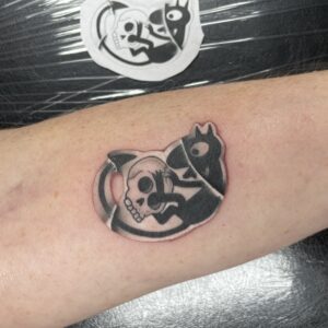 Forearm tattoo of a bold black-and-gray skull design with another dark figure overlapping it, partially visible on the skin.