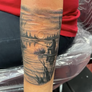 Forearm tattoo of a sunset over a lake with reeds, trees, and a small figure near the water on a dock-like platform
