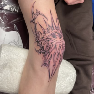 Fresh black outline tattoo on the inner forearm with an eye motif encircled by jagged, tribal-style spikes.