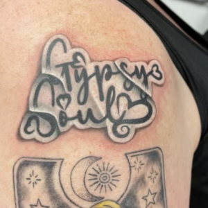 Close-up of a shoulder tattoo: cursive script with heart shapes above a framed portrait of a woman with blonde hair and celestial symbols (moon and stars).