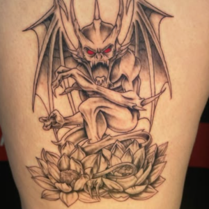 Tattoo of a menacing bat-demon with red eyes and spread wings perched on a lotus flower on the leg.