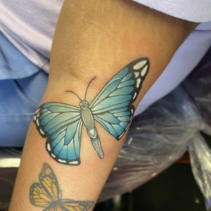 Blue butterfly tattoo on forearm with teal wings and black edges; smaller orange butterfly below it.