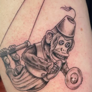 Tattoo of a cartoon monkey wearing a fez, gripping a long spear-like weapon and a round shield, all in black ink on the arm.