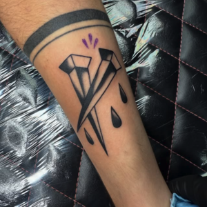 Geometric diamond tattoo on a forearm with three teardrop shapes beneath and a small purple accent above the diamond.