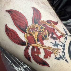 Colorful tattoo of a stylized red and yellow demon-insect creature with large wings and sharp skeletal limbs on a person's leg.
