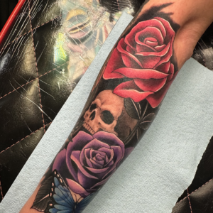 Color tattoo on forearm featuring red and purple roses, a skull, and a blue butterfly with dark shading in the background
