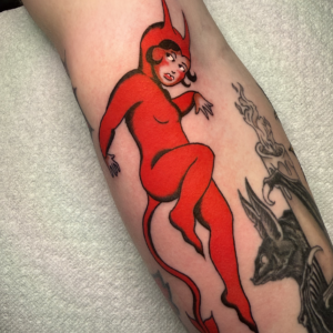 Red devil woman tattoo with horns and a long curling tail on the leg, posed as if dancing or floating forward.