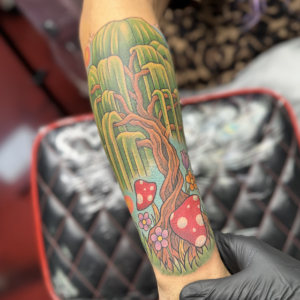 Forearm tattoo of a whimsical forest with twisting brown trunk, green trees, red spotted mushrooms and small flowers on skin.