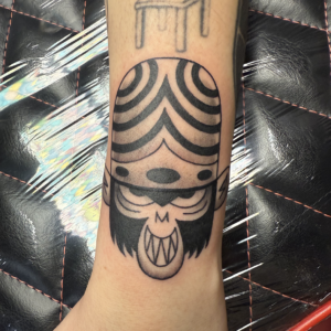 Tattoo on the forearm of a cartoon creature with striped helmet, big eyes, and a fanged mouth.