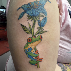 Colorful tattoo of a green frog clinging to a plant stem beneath a blue lily flower with orange stamens and leaves.