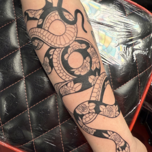Long black snake tattoo coiling around the forearm with rose patterns and scales, wrapping from wrist toward elbow.