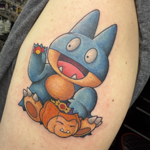 Colorful tattoo of a blue dragon-like creature with an orange belly sitting with a smiling orange pumpkin wearing a crown.